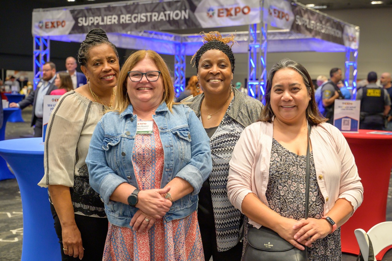 Four women smile and stand in front of the Supplier Registration booth.