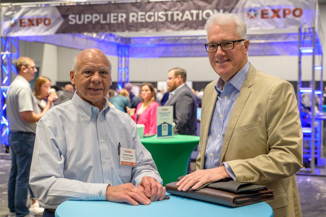 CharlesOrtega and an attendee smile at ballroom bistro table in front of the Supplier Registration booth.