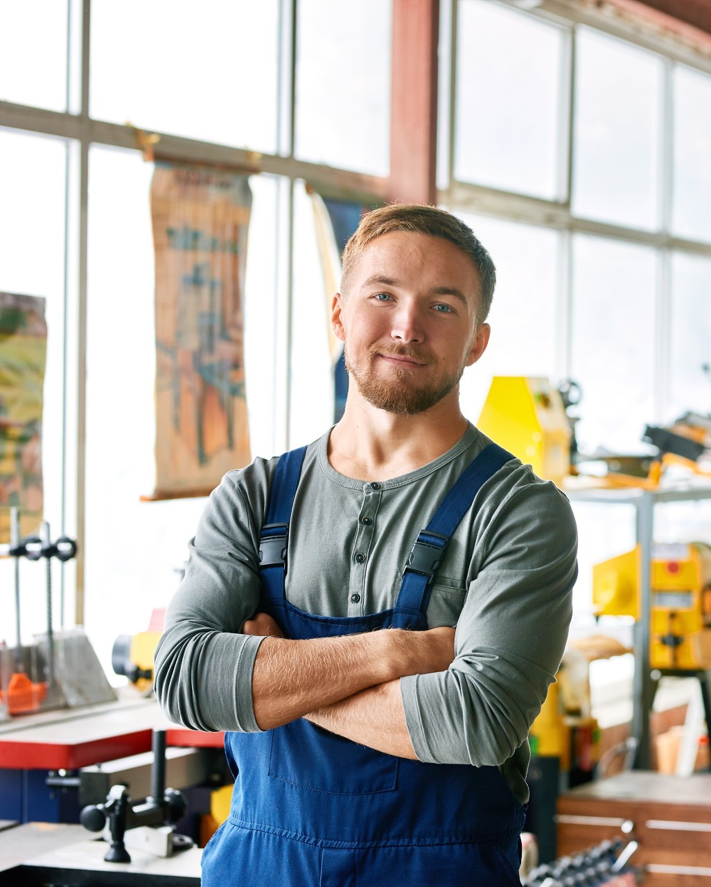 Portrait of smiling young man wearing overalls posing confidently with arms crossed at industrial factory with machine units, copy space