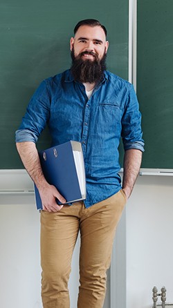 Relaxed confident male teacher in a classroom standing holding a binder in front of the chalkboard smiling at the camera
