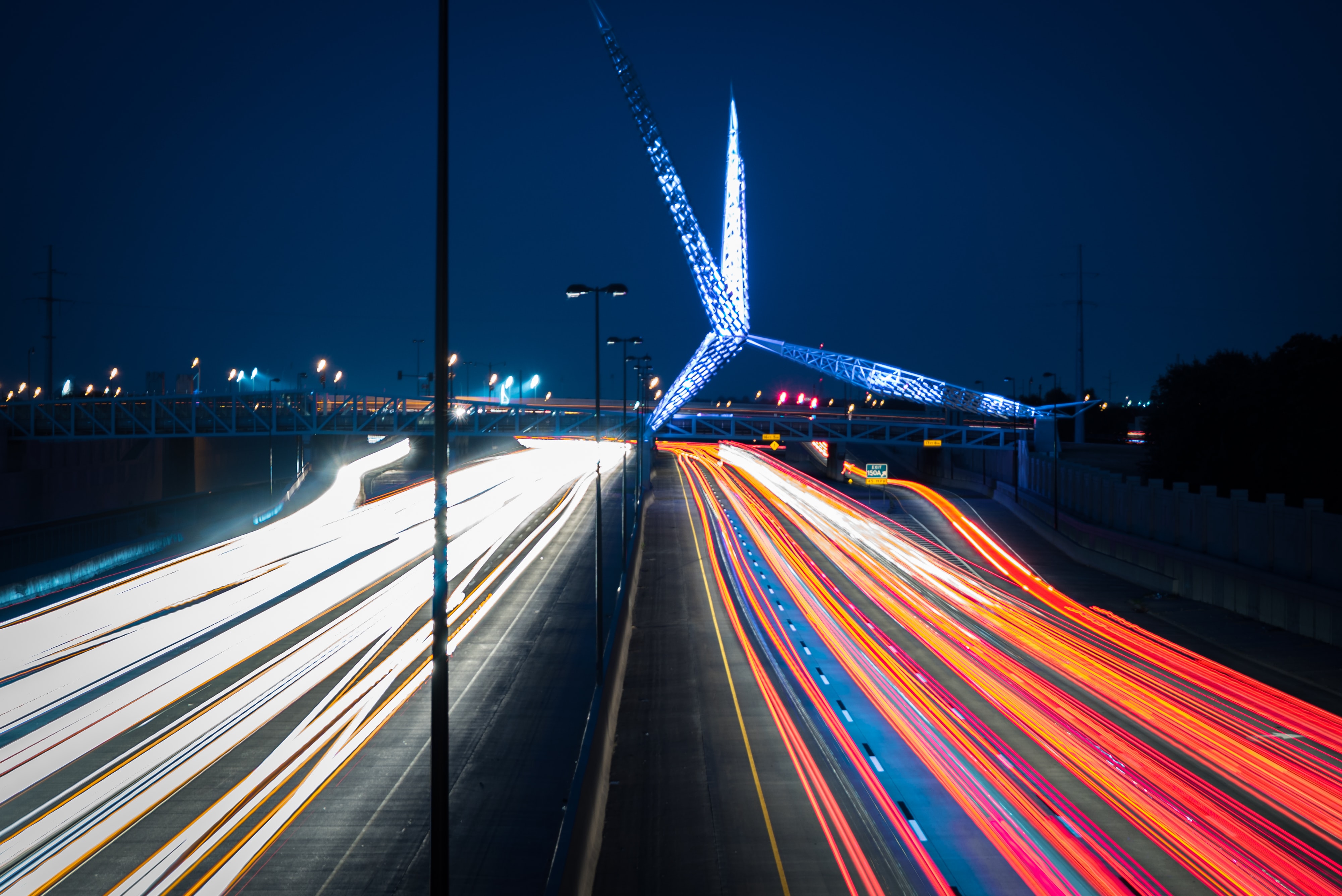 Blured cars driving at night under a blue lit Scissortail Bridge in downtown Oklahoma City