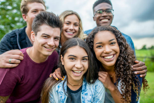 A diverse group of teens smiles with the arms around each other's shoulders, positing for a group photo. They are outside and there is a green meadow behind them.