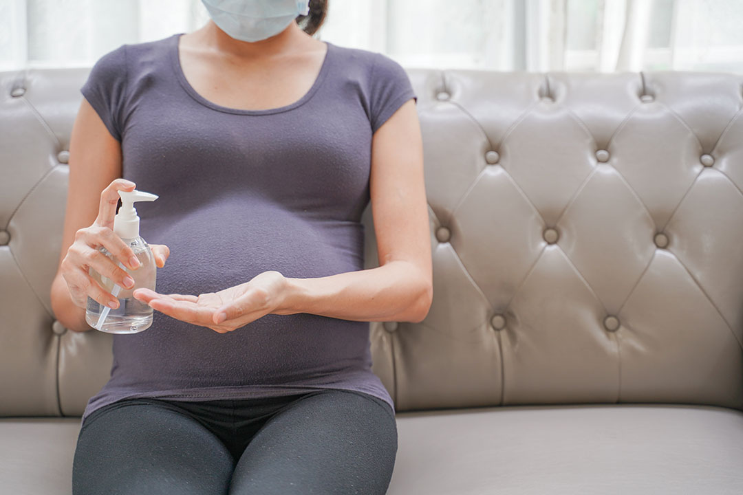 A pregnant woman wears a surgical mask is washing hands by alcohol gel on the sofa. Protect a COVID-19 (Coronavirus), PM 2.5 and prevent infection to the fetus concept.