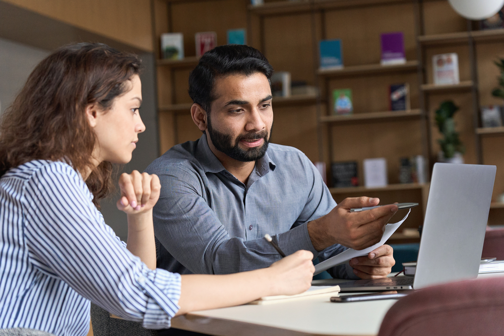 Professional indian teacher, executive or mentor helping latin student, new employee, teaching intern, explaining online job using laptop computer, talking, having teamwork discussion in office.