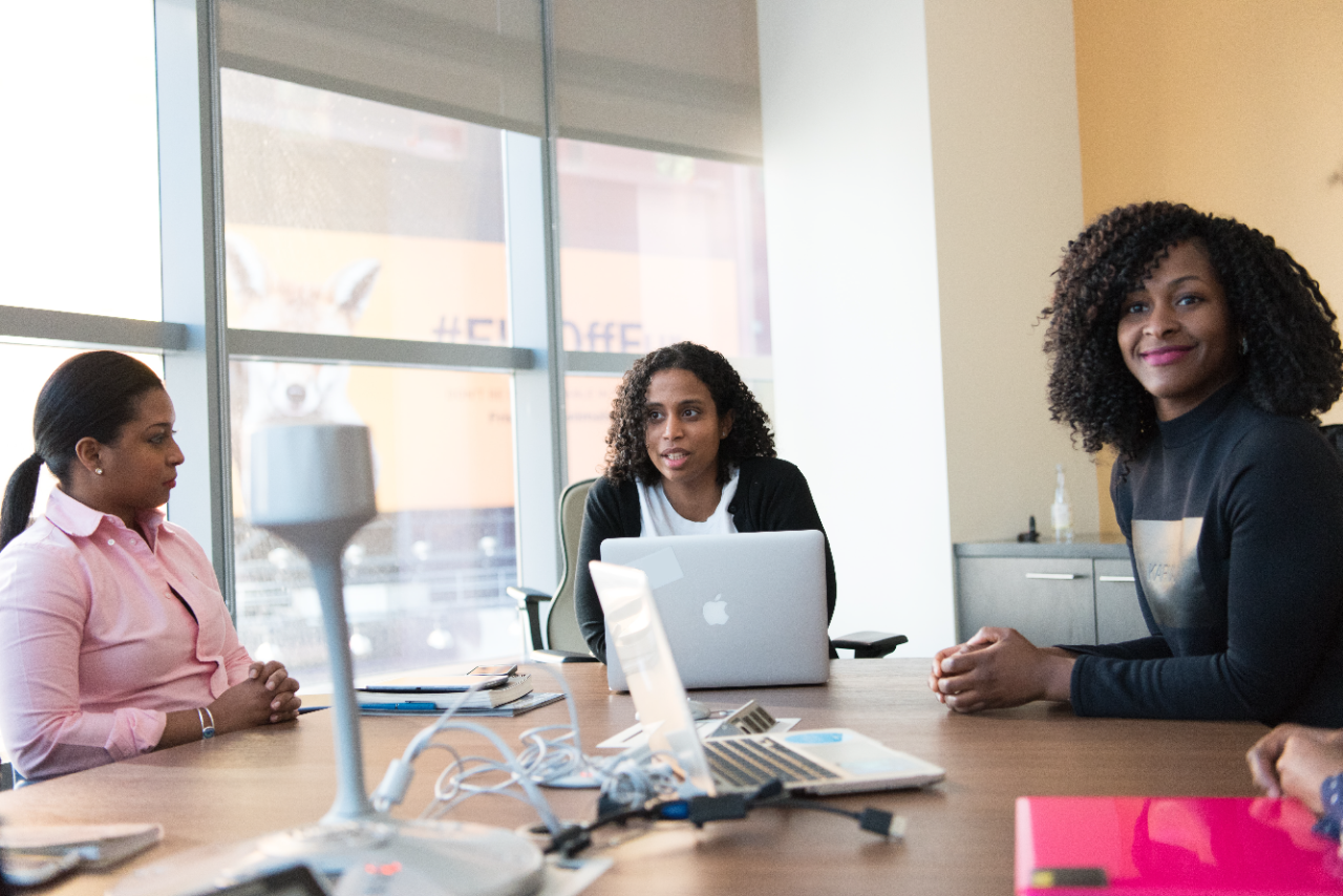 Four Women At Meeting Table with windows in background and three laptops on the table