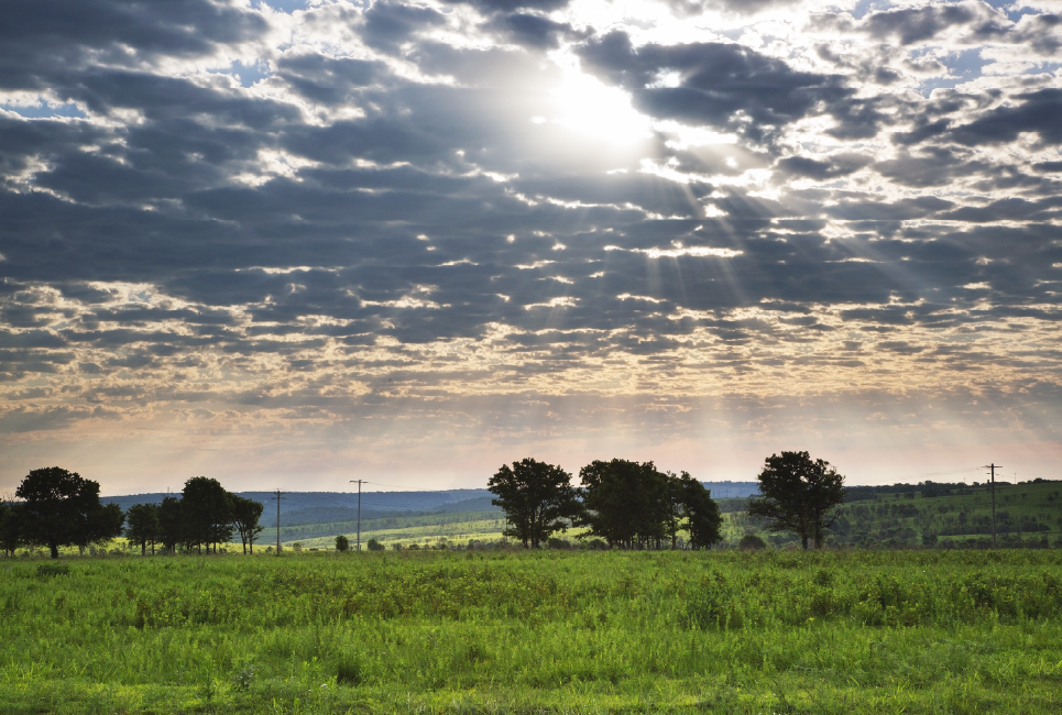 oklahoma prairie with green fields, a few trees, and the sun peeking through mostly-cloudy skies