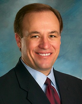 Head shot of Todd Hiett smiling wearing black suit and red tie in front of blue marbled background