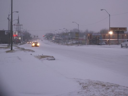 Dusk snow cover street with cars with their lights on. 