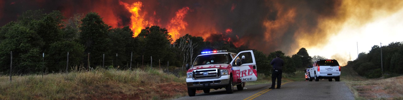 A fire truck on the side of the road and another is blocking the road with a wildfire burning in behind them.