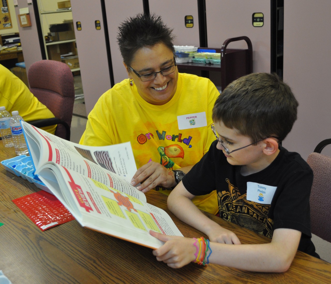 A woman sits a table with a young boy, together they are reading a large print book.