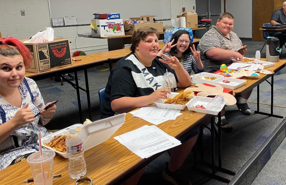 Students smiling and making peace signs in a classroom with lunch and paperwork on the table. 